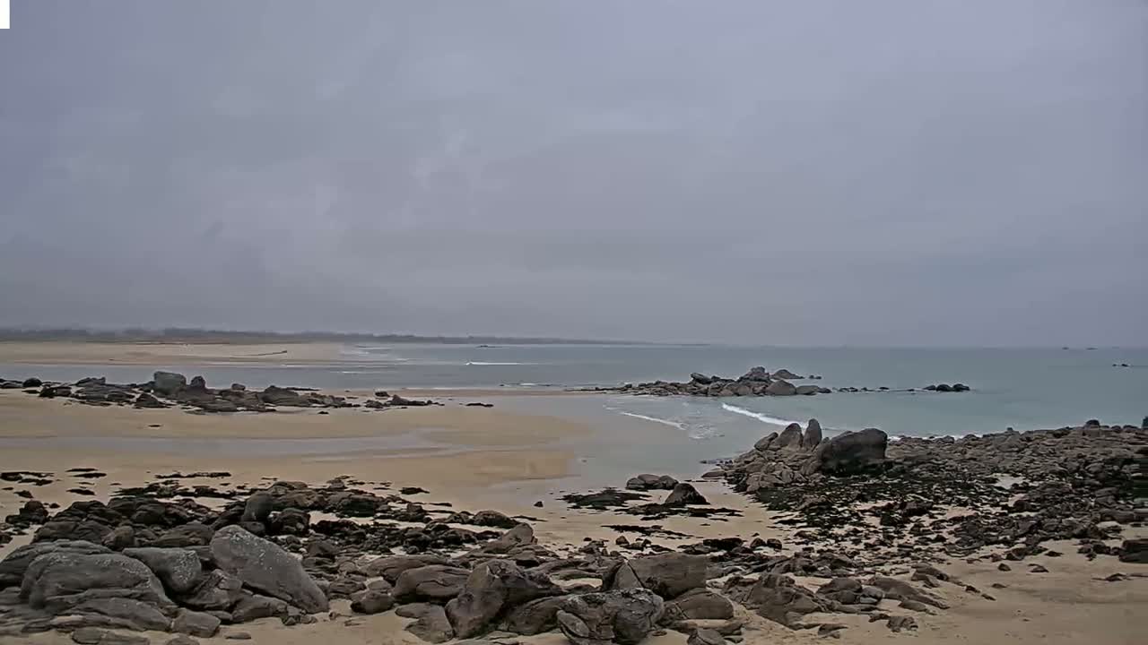 A sandy beach with dark rocks and a calm, light-blue ocean under an overcast sky.