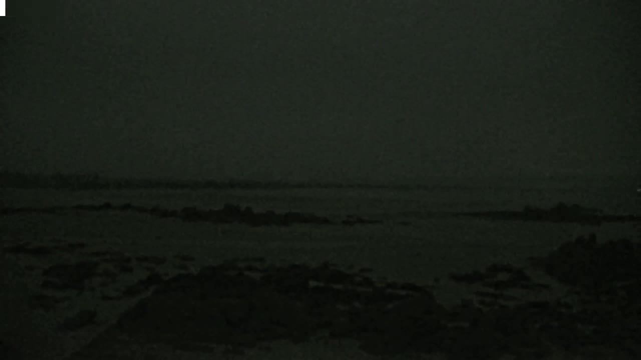A dark, overcast sky hangs over a rocky shoreline and calm body of water.