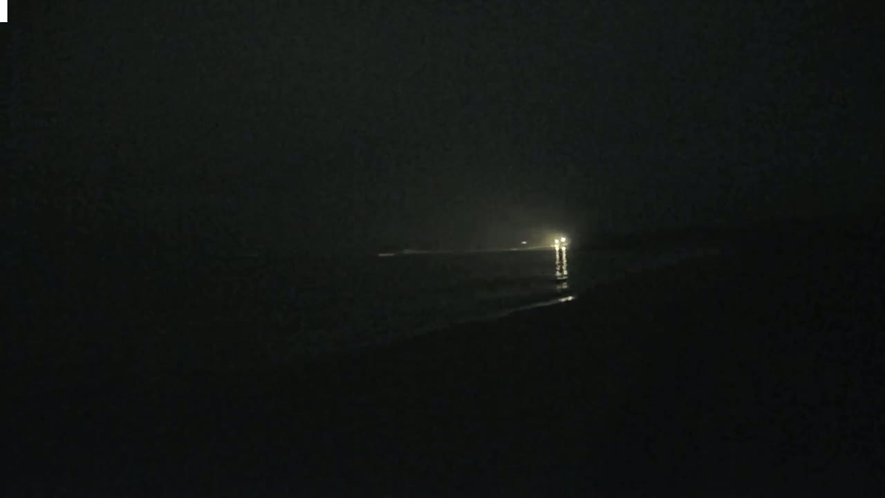 A dimly lit, nighttime beach scene shows a faint light source on the water's edge in the distance.