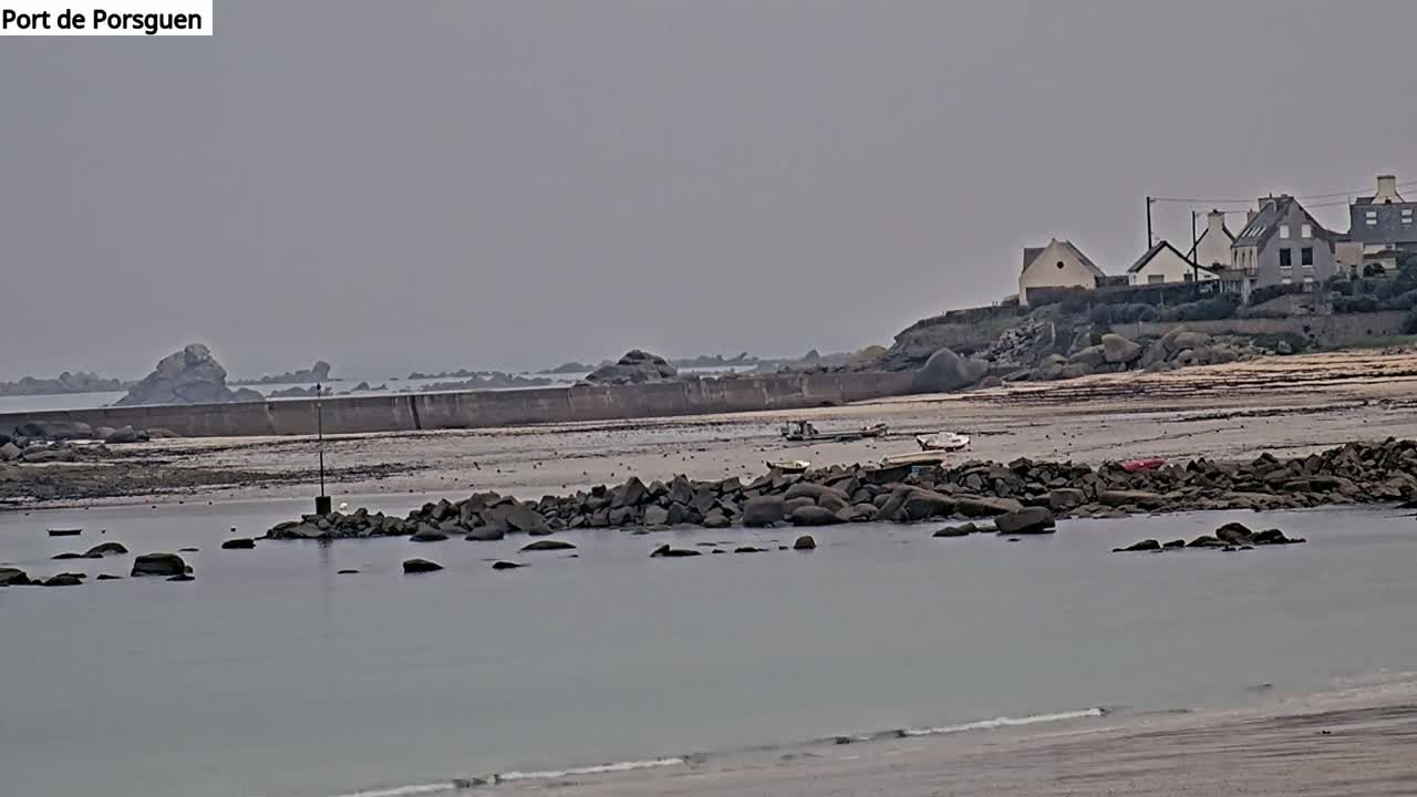 A calm, overcast day reveals a rocky shoreline with a low seawall, several small boats, and a cluster of buildings on a hill in the distance.