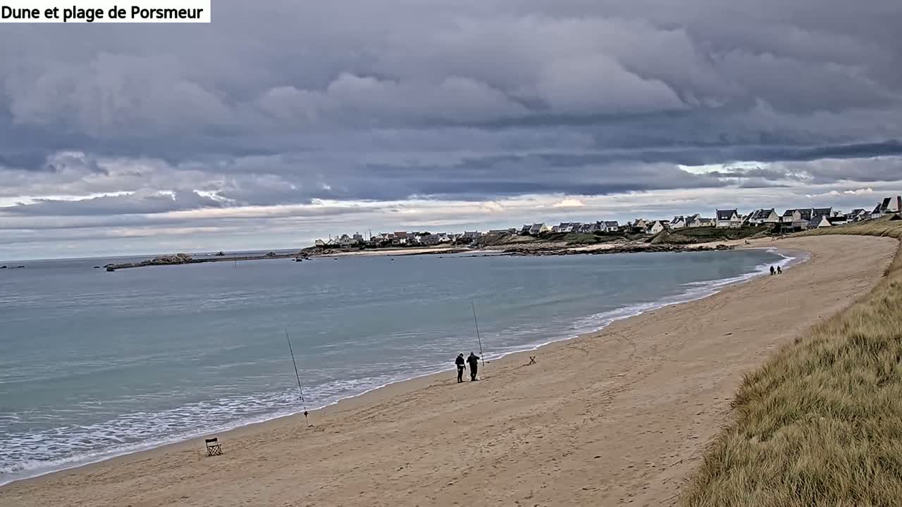 A sandy beach curves gently around a calm bay under a cloudy sky, with two people fishing along the shoreline and a small village visible in the distance.