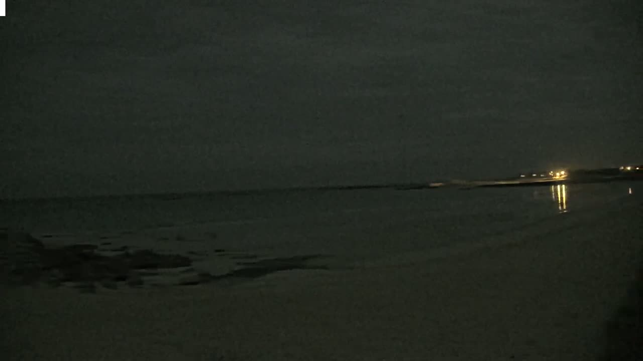 A dark, nighttime beach scene shows a calm ocean with faint lights reflecting on the water from a distant shore under a cloudy sky.
