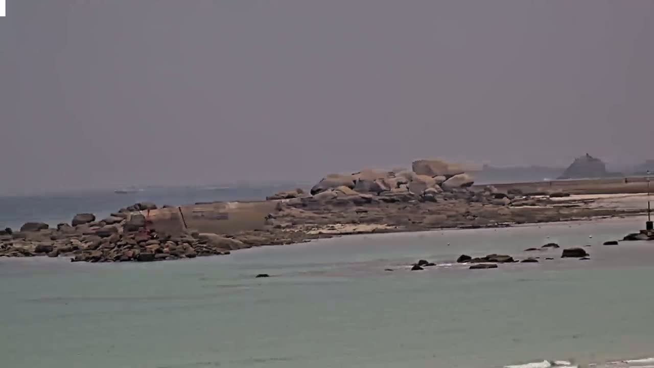 A hazy, overcast day reveals a rocky shoreline with a low-lying stone structure and calm, light-green water.