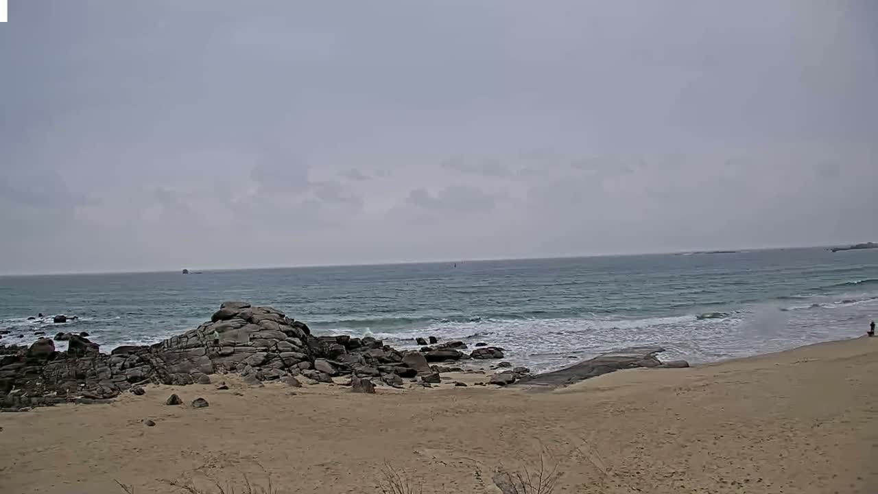 A sandy beach with dark rocks meets a somewhat choppy sea under a cloudy sky.