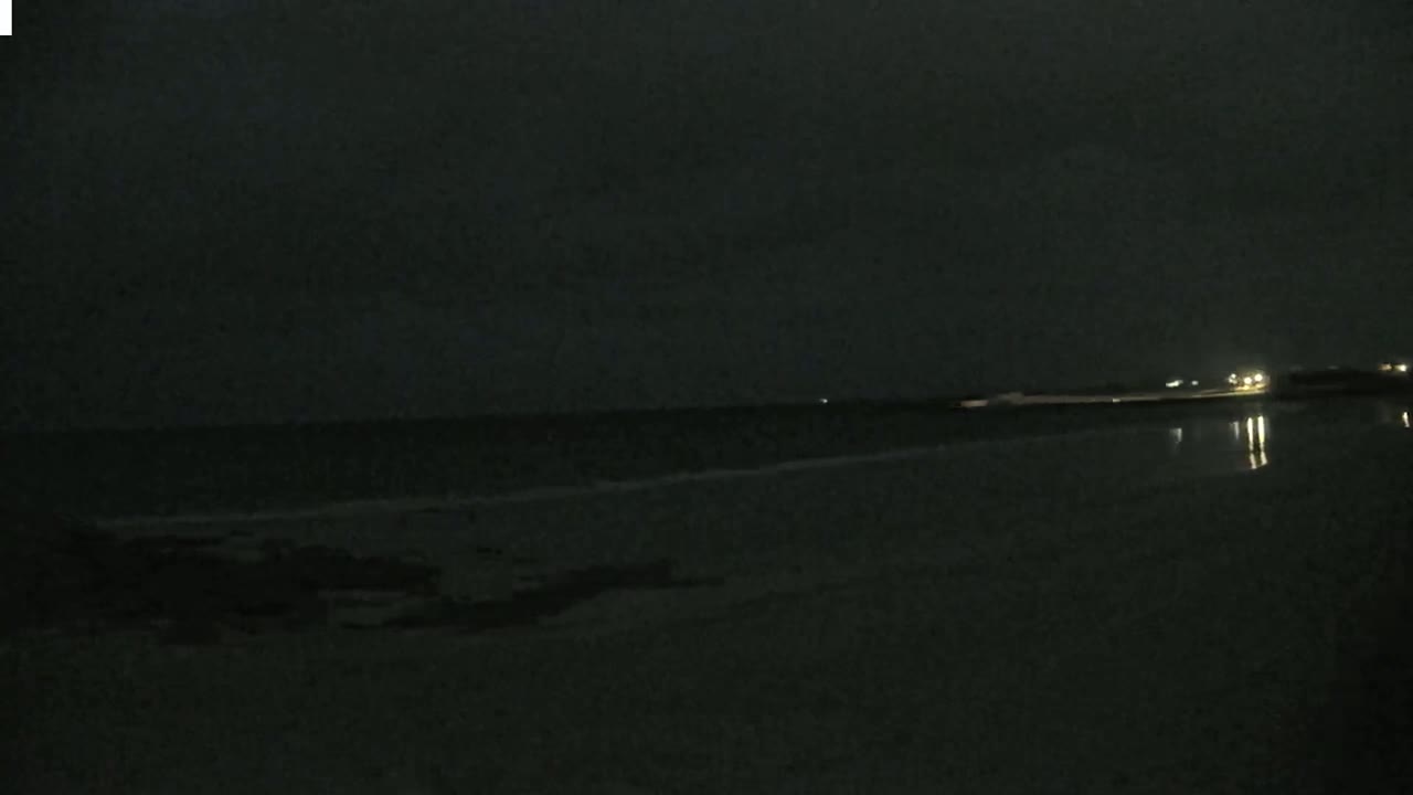 A dark, nighttime scene shows a calm body of water reflecting a few distant lights under a cloudy sky.