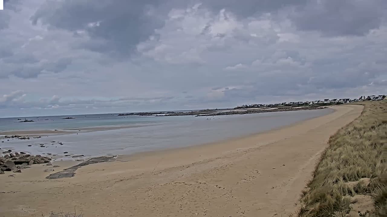 A sandy beach stretches towards a calm sea under a cloudy sky, with a line of houses visible in the distance.