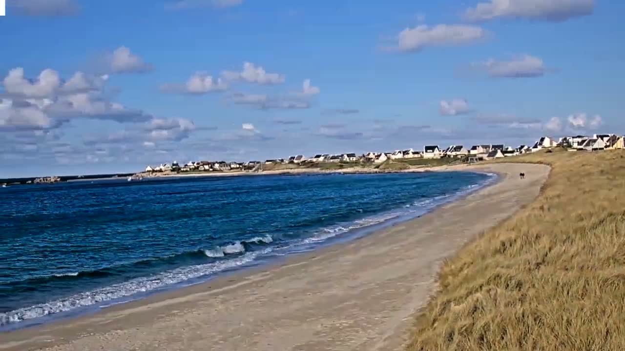 A sandy beach curves gently around a calm, blue ocean under a partly cloudy sky, with a small village visible in the distance.