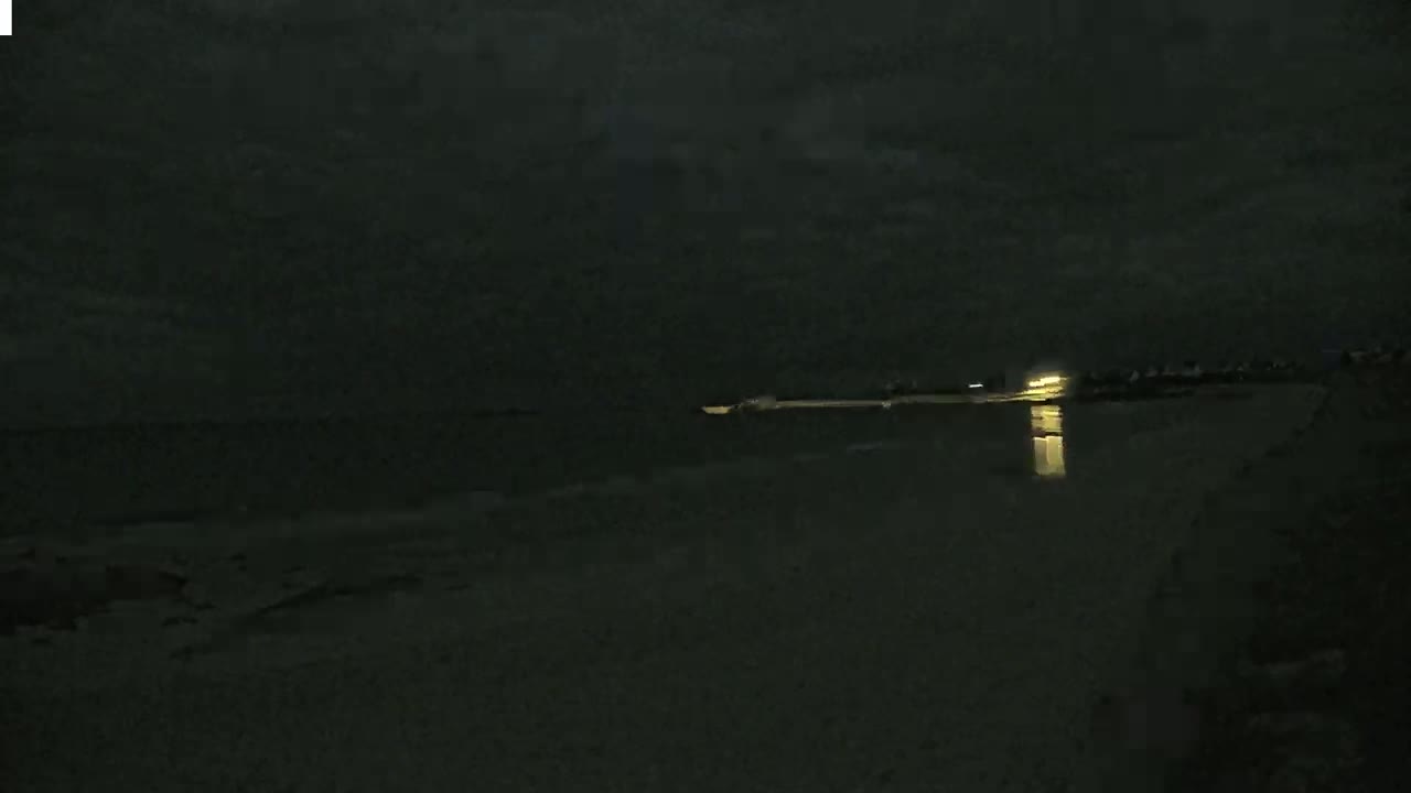 A dark, nighttime beach scene shows a calm body of water reflecting a distant light source under a cloudy sky.