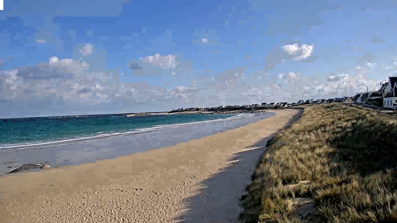 A sandy beach stretches along a turquoise ocean under a partly cloudy blue sky, with houses visible in the distance.
