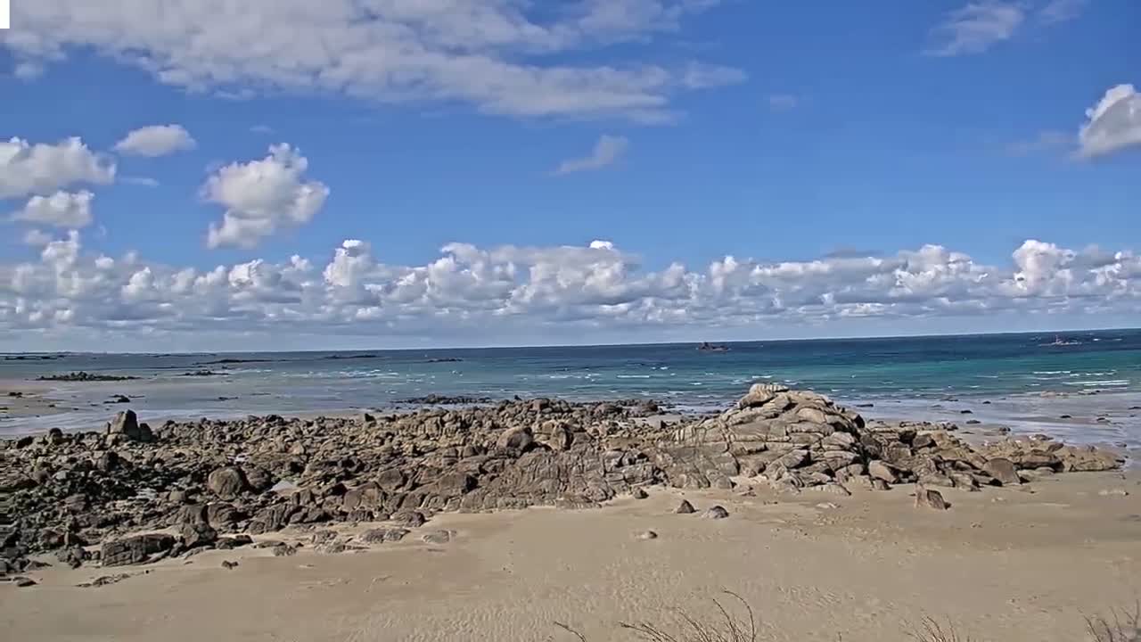 A sandy beach with dark rocks stretches to a calm, turquoise ocean under a partly cloudy blue sky.
