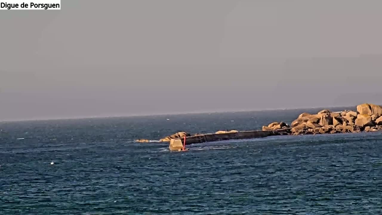 A rocky outcrop connected to a seawall by a low-lying structure extends into a calm, dark-blue sea under a hazy sky.