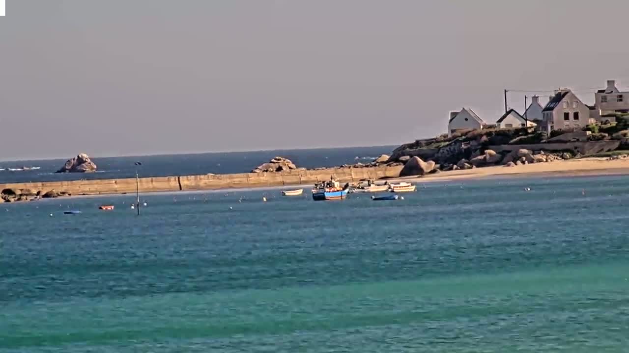 Several small boats are moored in a calm, teal-colored bay near a rocky shoreline with a few houses on a hill.