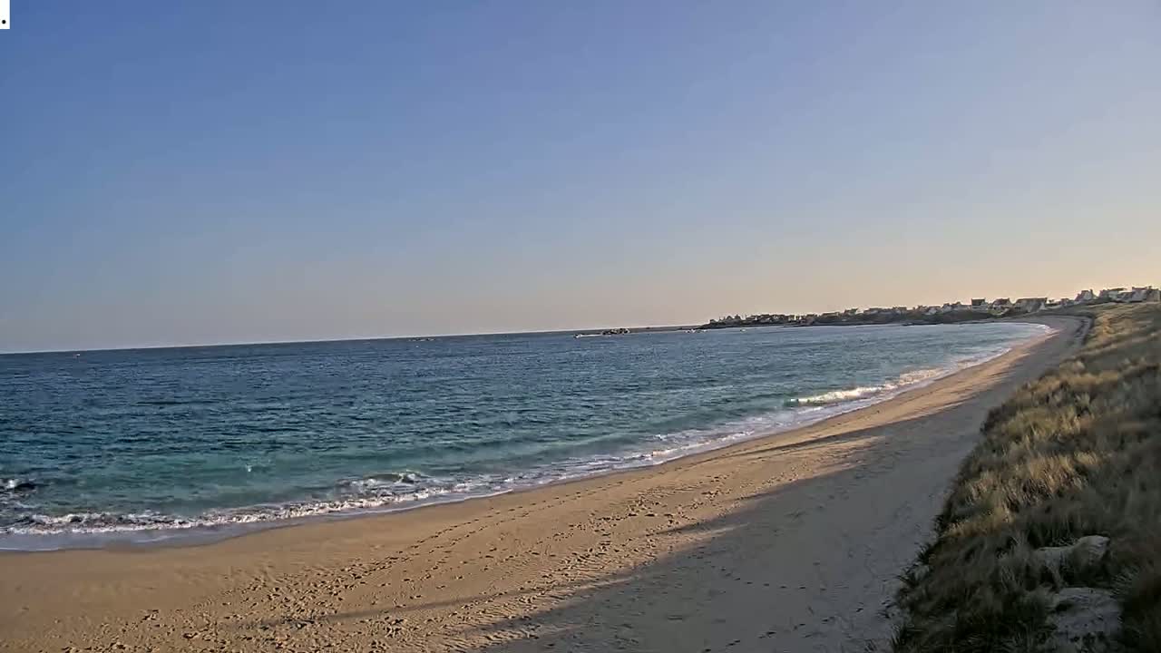A sandy beach curves gently to the right, meeting a calm, light-blue ocean under a clear, sunny sky.