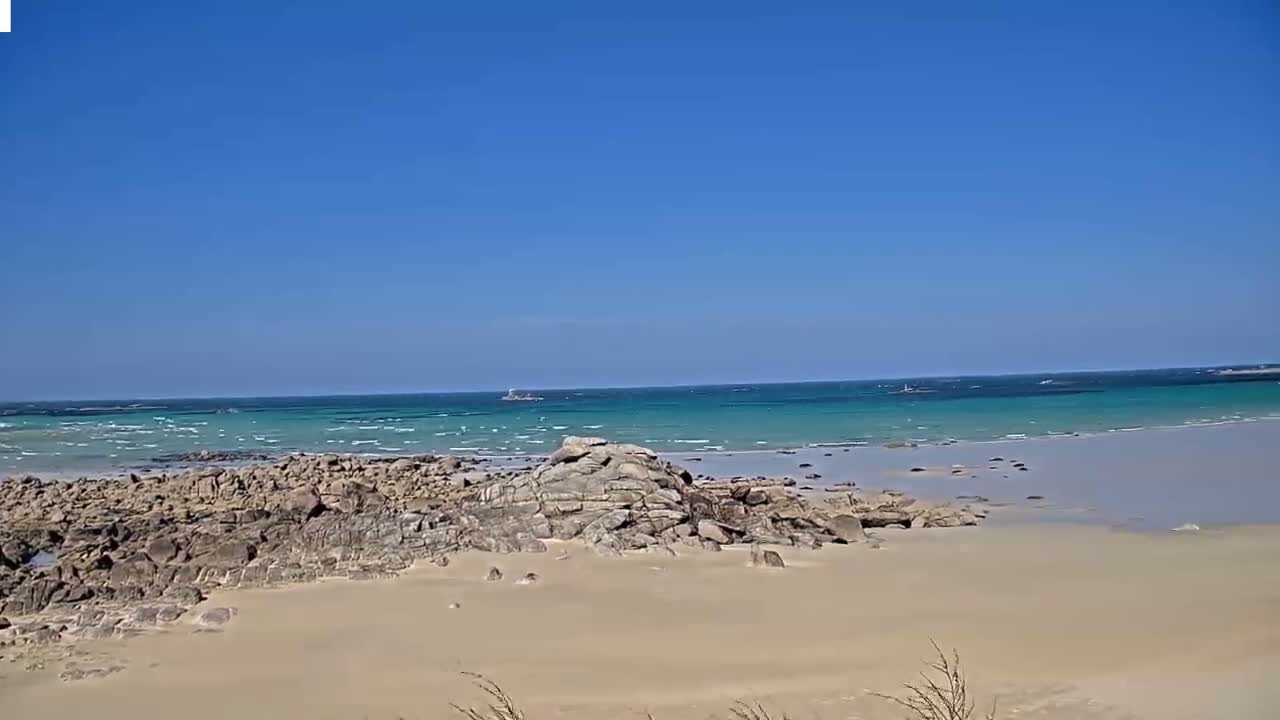 A sandy beach with dark rocks stretches toward a calm, teal ocean under a clear blue sky.
