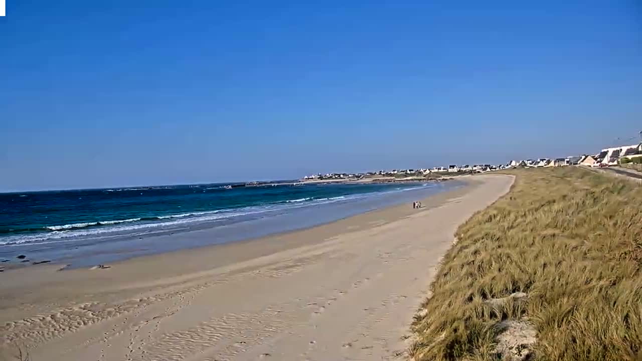 A sandy beach with gentle waves under a clear blue sky stretches towards a small town in the distance.