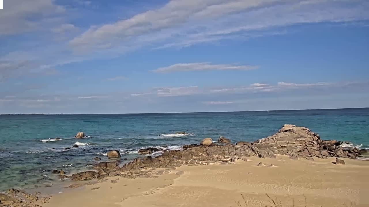 A sandy beach with dark rocks and small waves laps the shore under a partly cloudy blue sky.