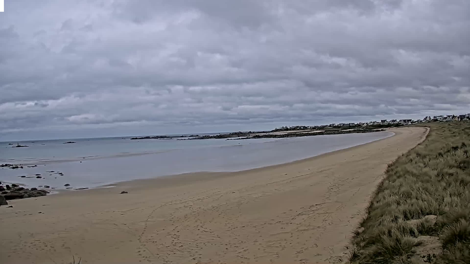 A sandy beach curves along a calm, gray ocean under a cloudy sky.