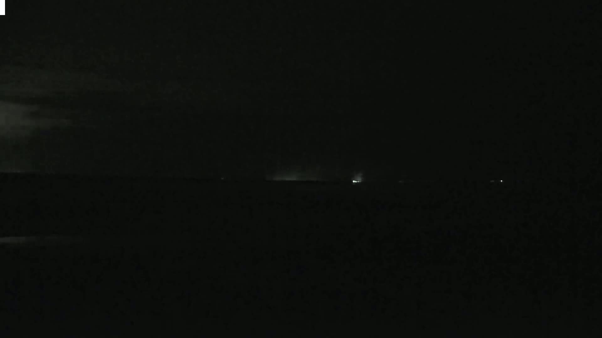 A dark, nighttime landscape shows a faint horizon line with a few distant lights under a cloudy sky.