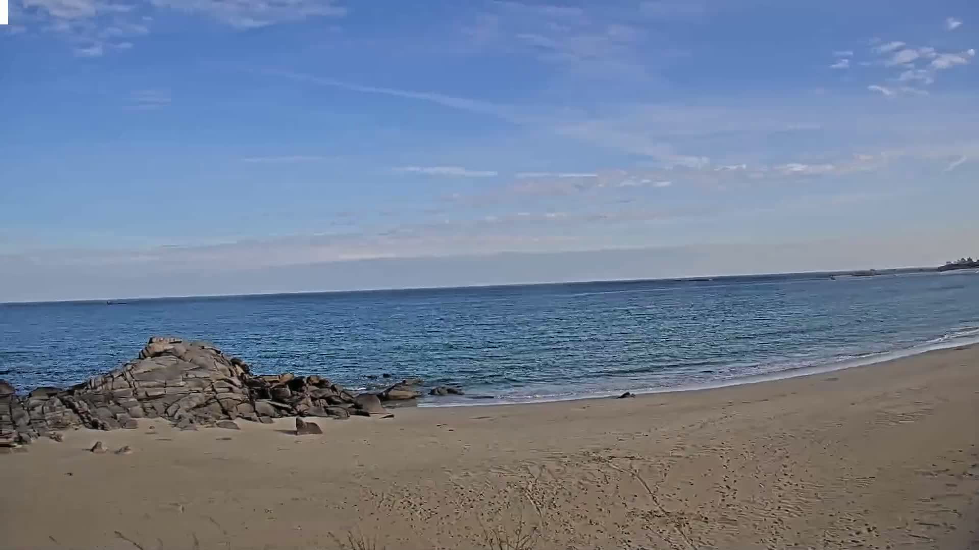 A sandy beach curves gently around dark rocks meeting a calm, blue ocean under a mostly sunny sky.