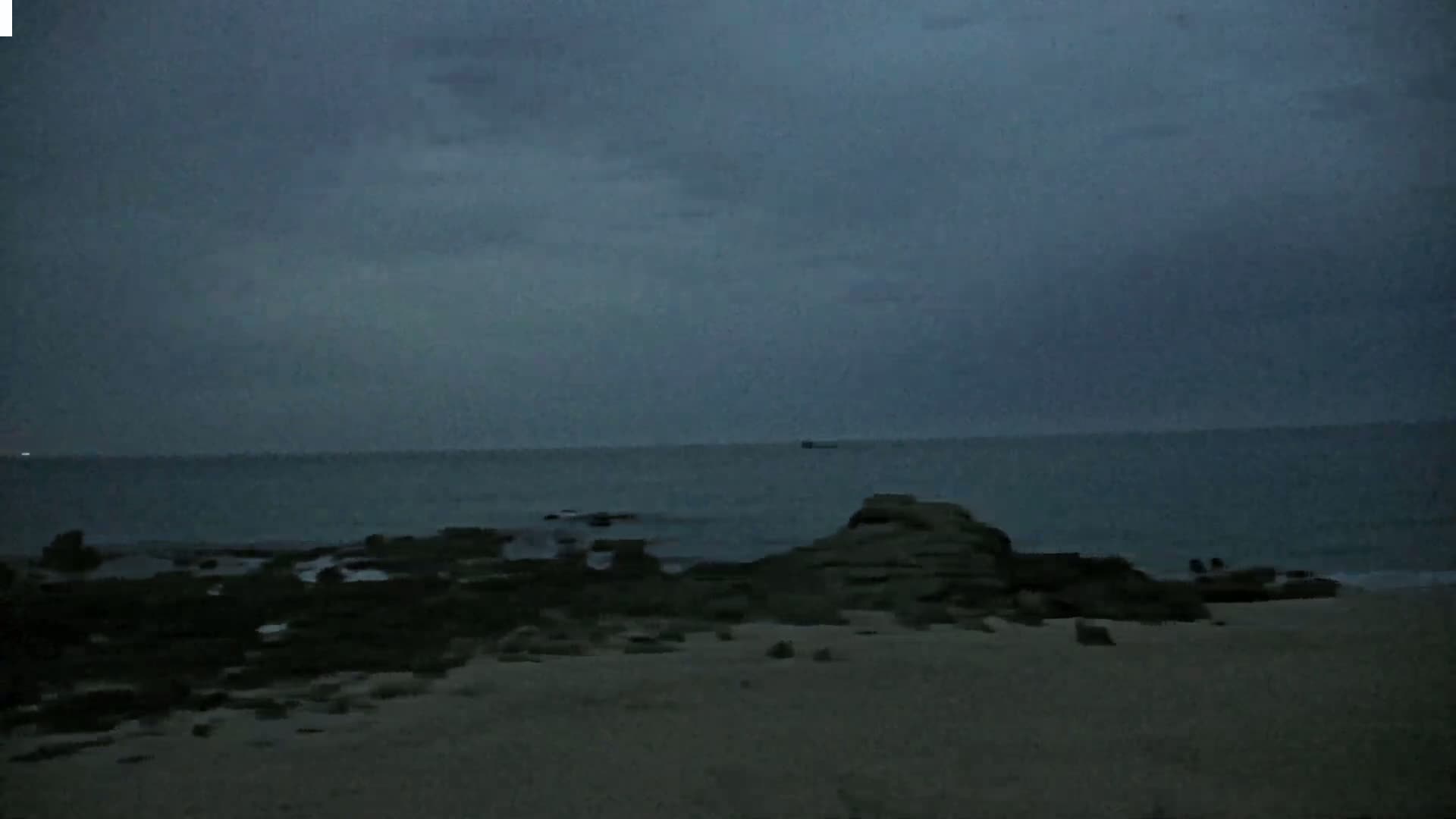 A dark, overcast sky hangs over a calm ocean, where dark rocks meet a sandy beach.