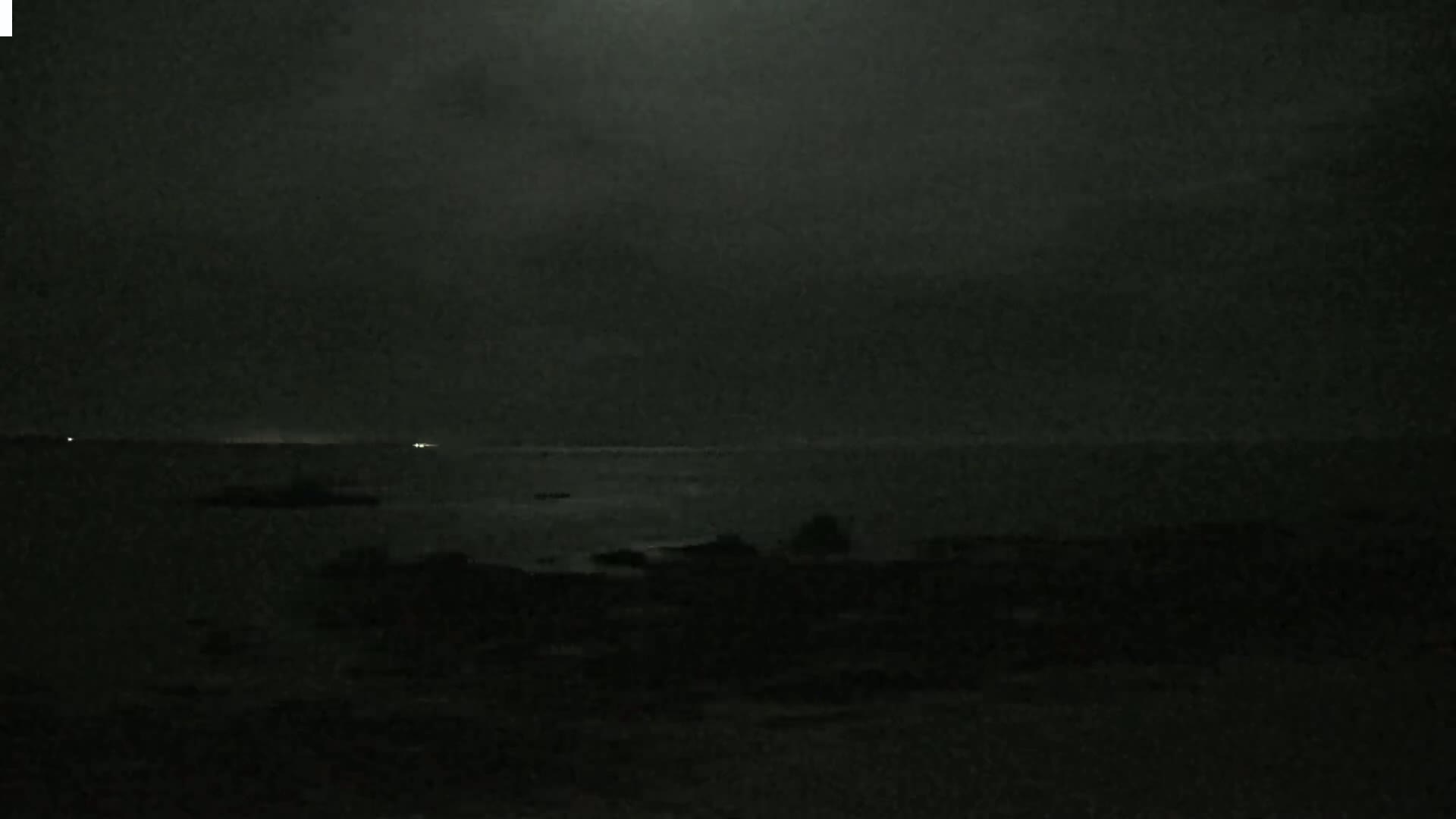 A dark, nighttime scene shows a calm body of water under a cloudy sky with faint lights visible on the horizon.