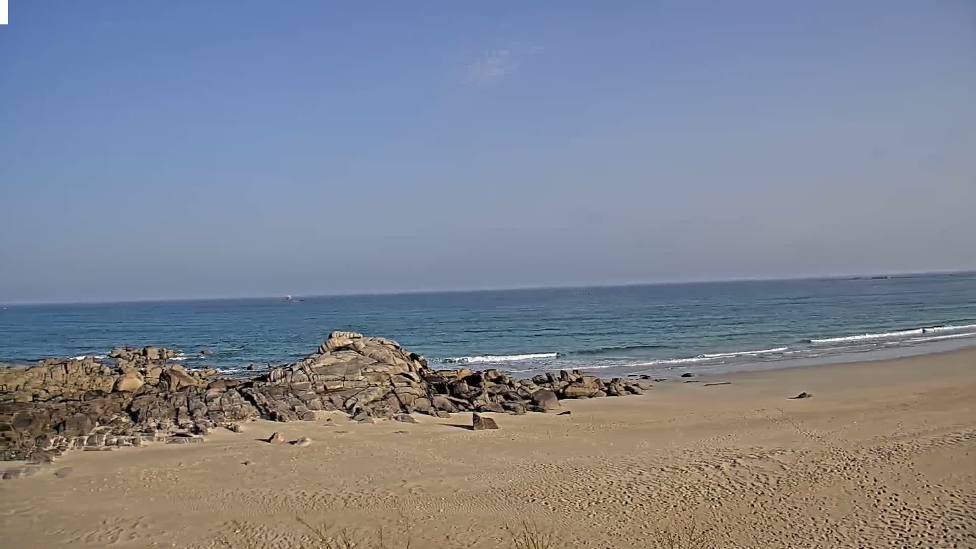 A sandy beach with dark rocks near the water's edge, under a clear blue sky, features gently rolling waves on a calm ocean.