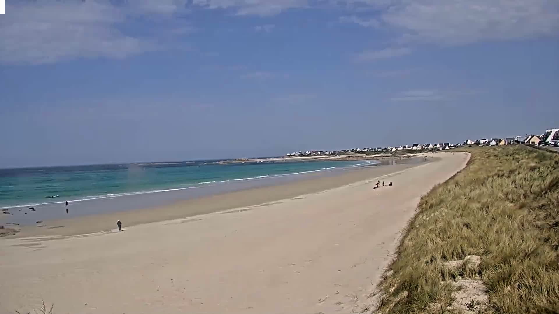 A long, sandy beach stretches out to a calm, turquoise ocean under a mostly sunny sky, with a few people visible on the shore and a small town in the distance.