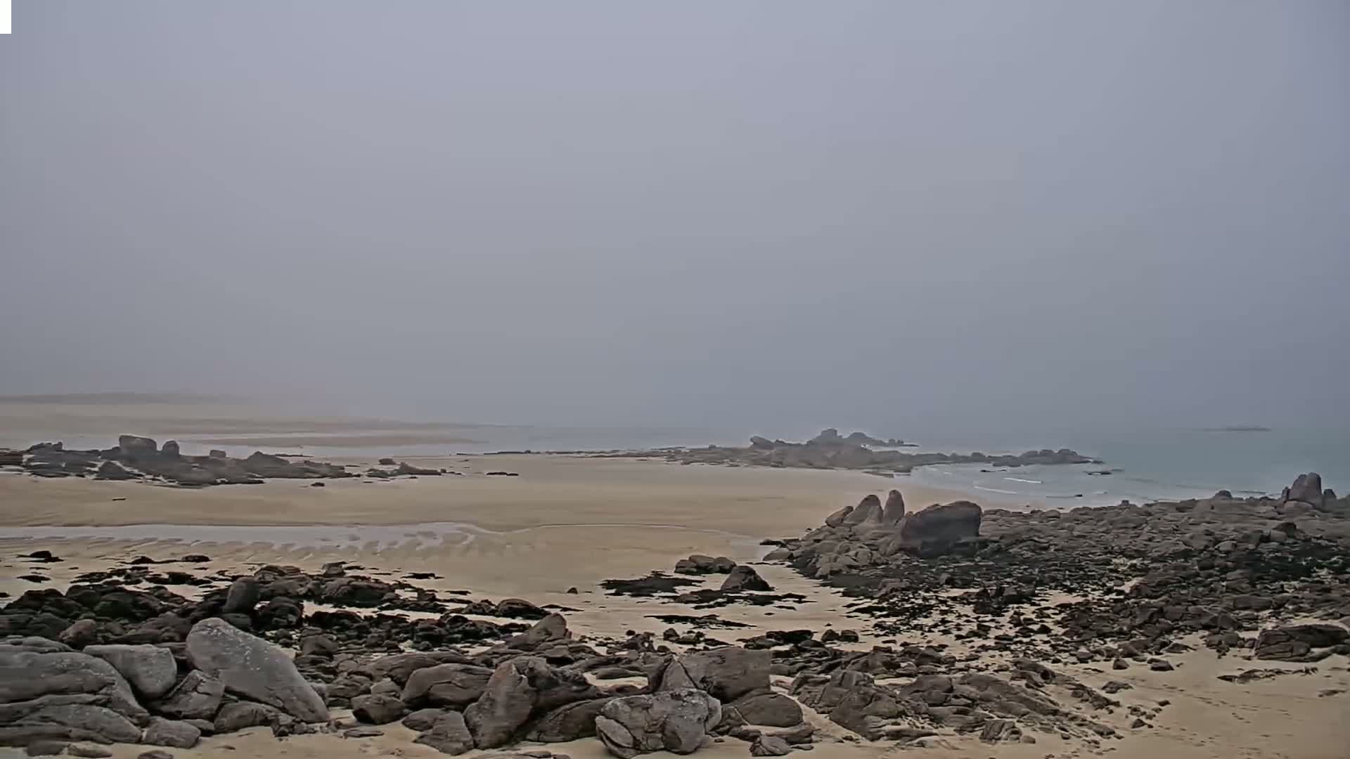 A sandy beach stretches to a hazy ocean, bordered by dark rocks under a foggy sky.