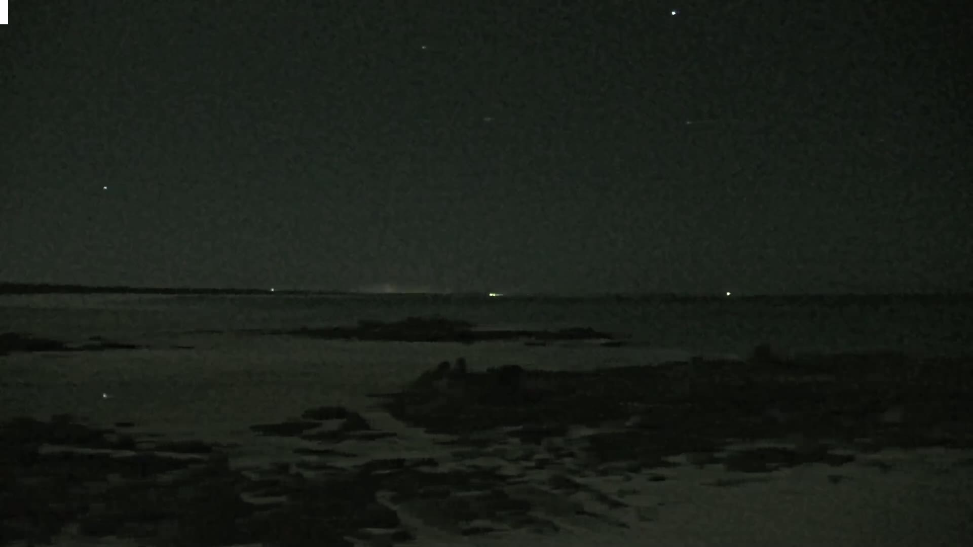 A dark, nighttime scene shows a calm body of water under a starlit sky, with a rocky shoreline in the foreground.