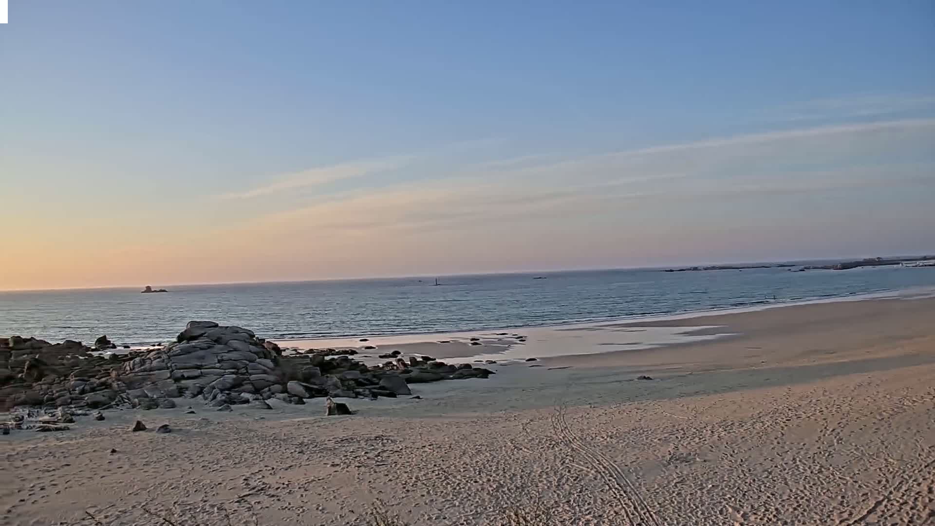 A sandy beach with dark rocks at the edge meets a calm ocean under a pale sunset sky.
