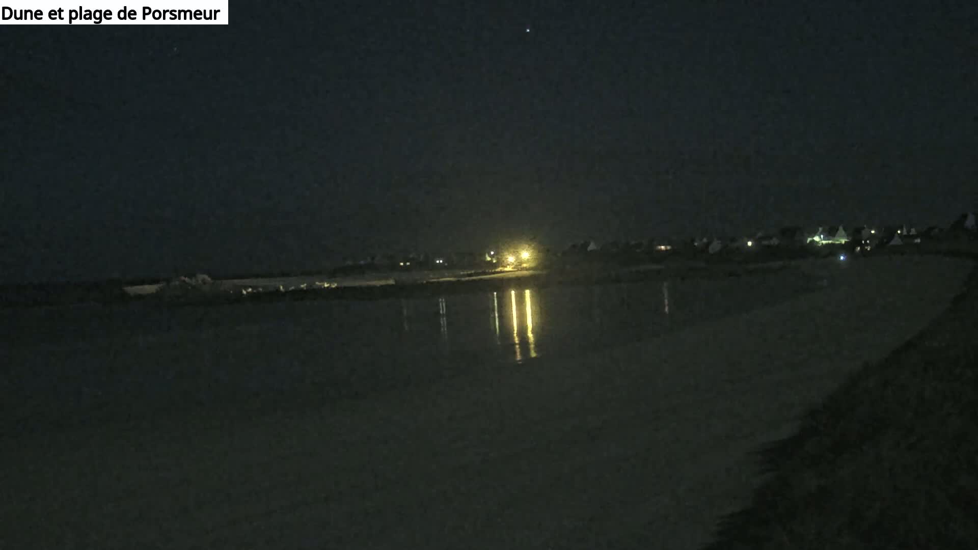 A dark, nighttime view of a calm body of water reflecting the lights of a distant town under a clear, starlit sky.