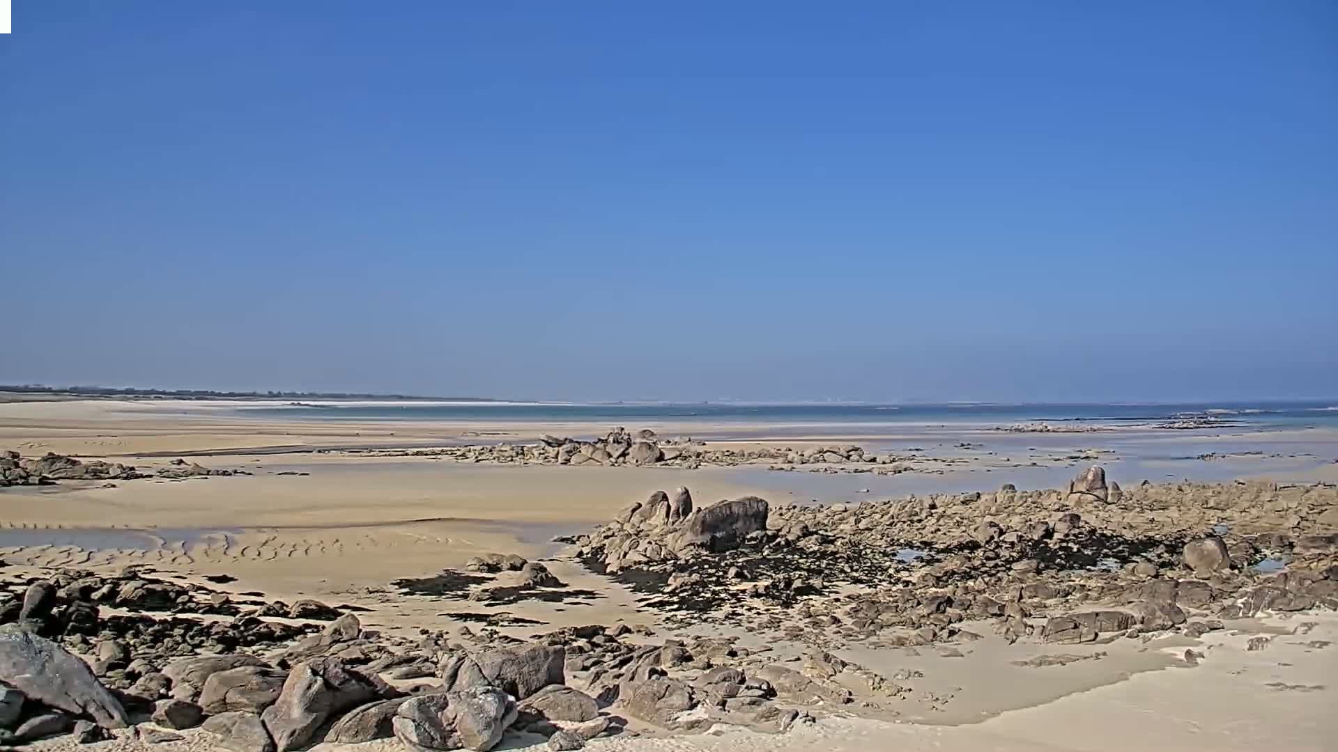 A sandy beach with dark rocks and shallow water stretches to a calm ocean under a clear blue sky.