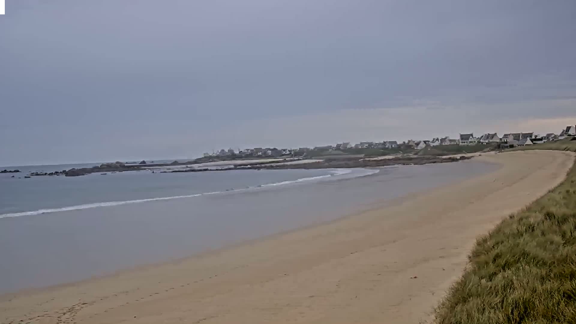 A sandy beach curves gently to the left, meeting calm, light-grey water under an overcast sky, with a small village visible in the distance.