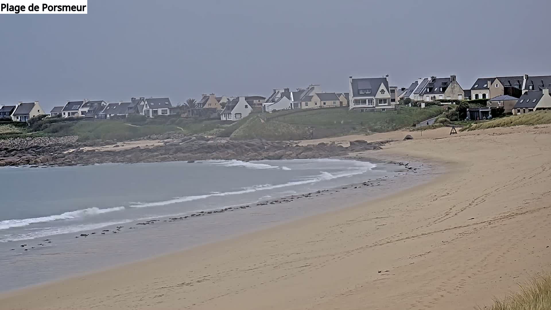 A sandy beach with gentle waves laps a shoreline of rocks, behind which sits a row of houses under an overcast sky.