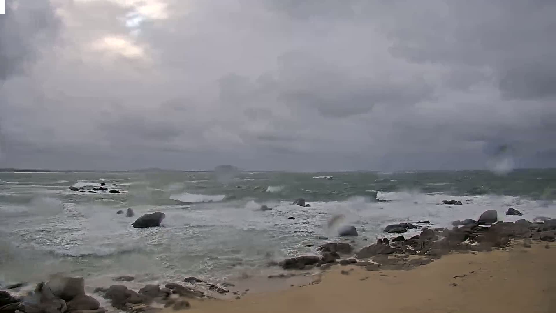 A rocky shoreline with a sandy beach is battered by a stormy sea under a gray, overcast sky.