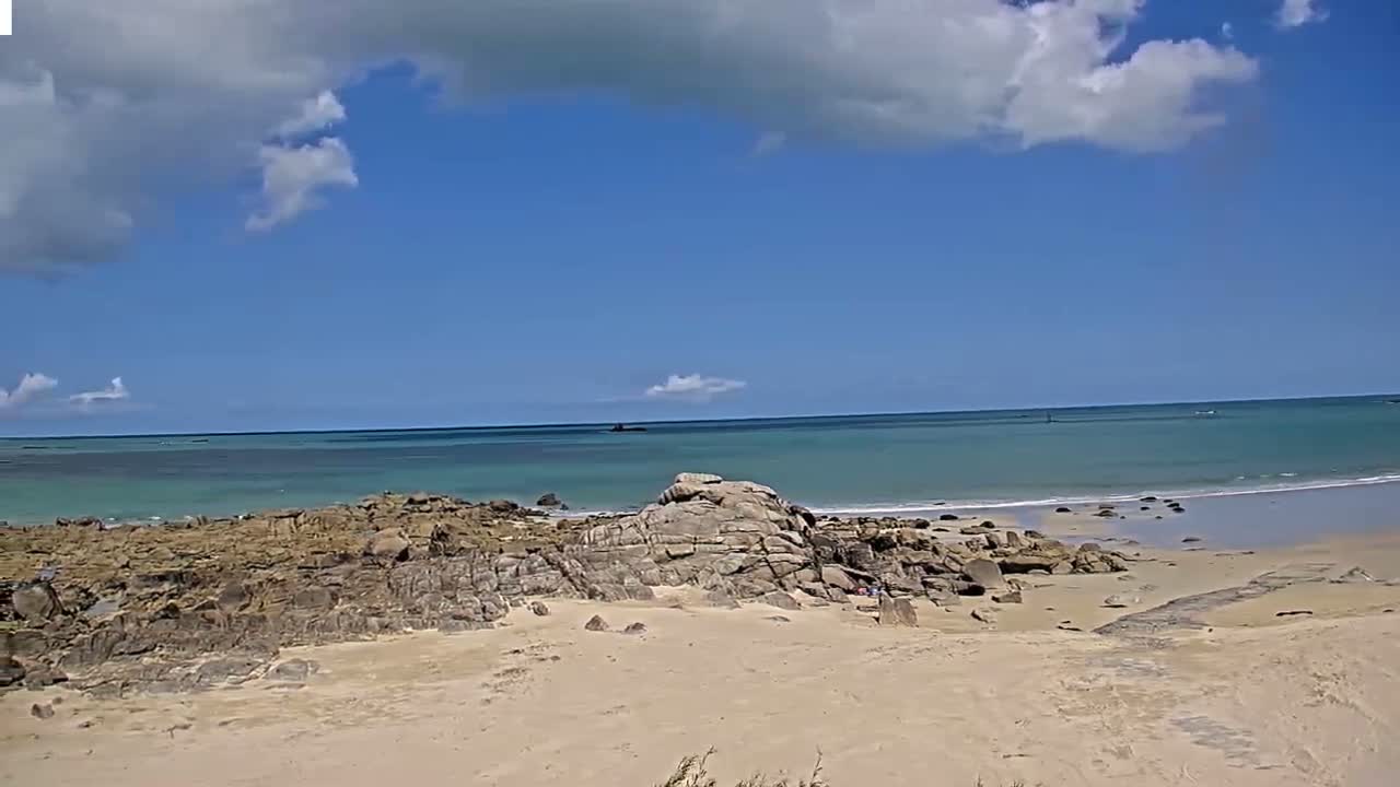 A sandy beach with rocky outcrops stretches to a calm, turquoise ocean under a partly cloudy sky.