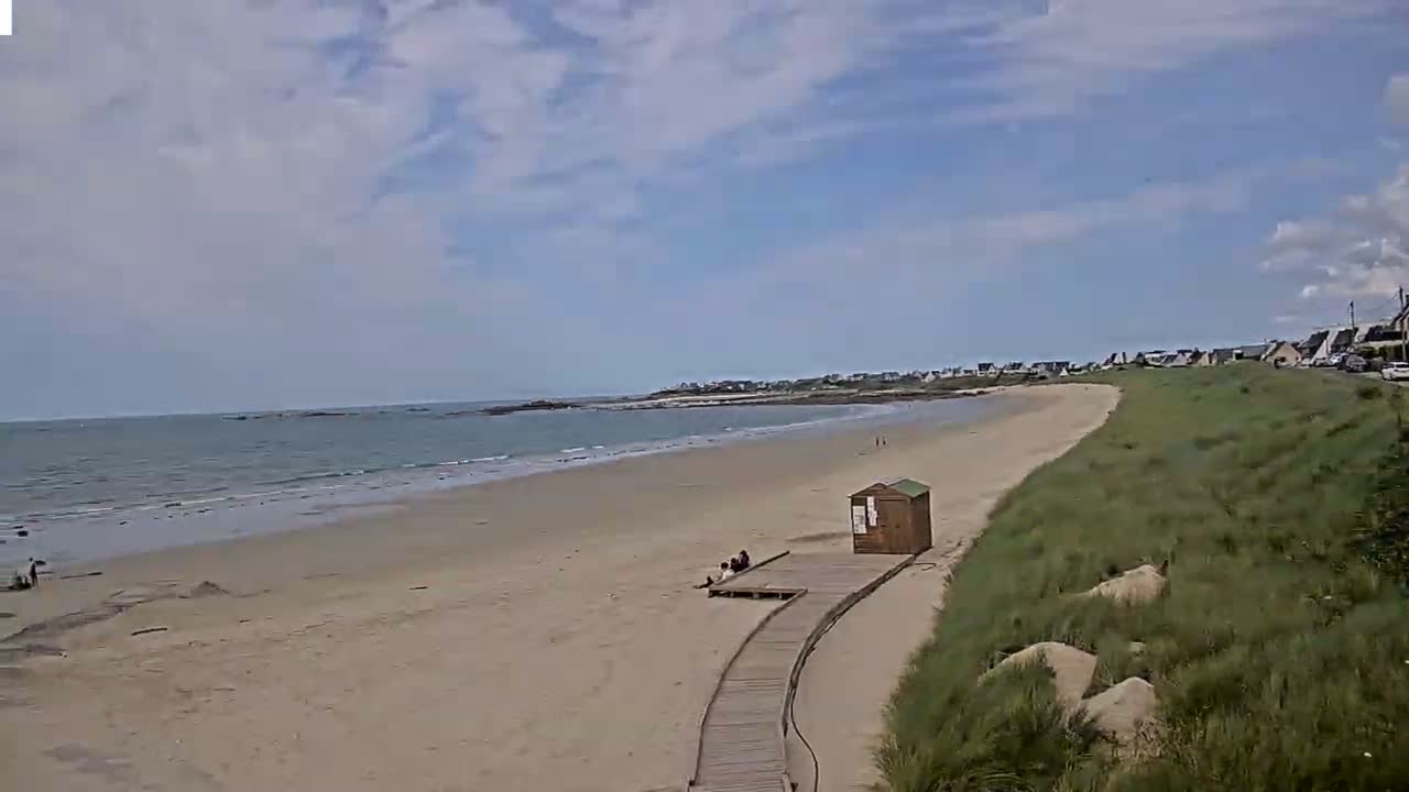 A sandy beach with calm waves stretches toward a distant shoreline under a partly cloudy sky, and a wooden boardwalk leads to a small structure on the sand.