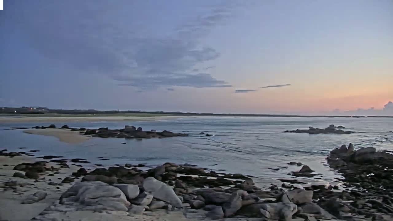 A sandy beach with calm waves stretches toward a distant shoreline under a partly cloudy sky, and a wooden boardwalk leads to a small structure on the sand.