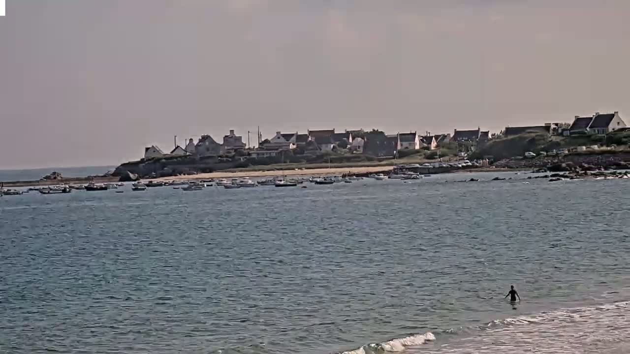 A small coastal town sits on a hill overlooking a calm bay with numerous small boats, and a person is wading in the shallow water of a nearby beach on a hazy day.