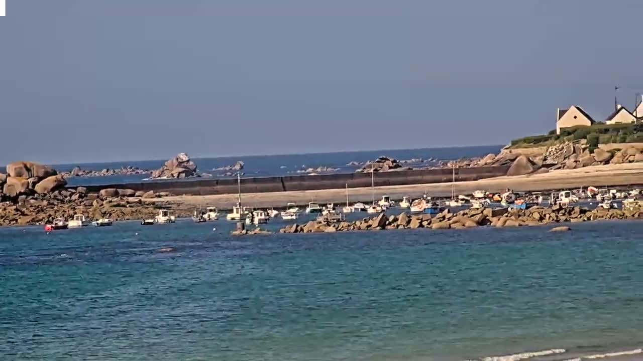 A calm, sunny day shows a small harbor filled with numerous boats nestled between rocky shores and a larger body of water.