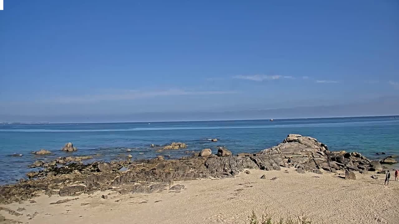 A sandy beach with dark rocks and a calm, clear blue ocean under a sunny sky.