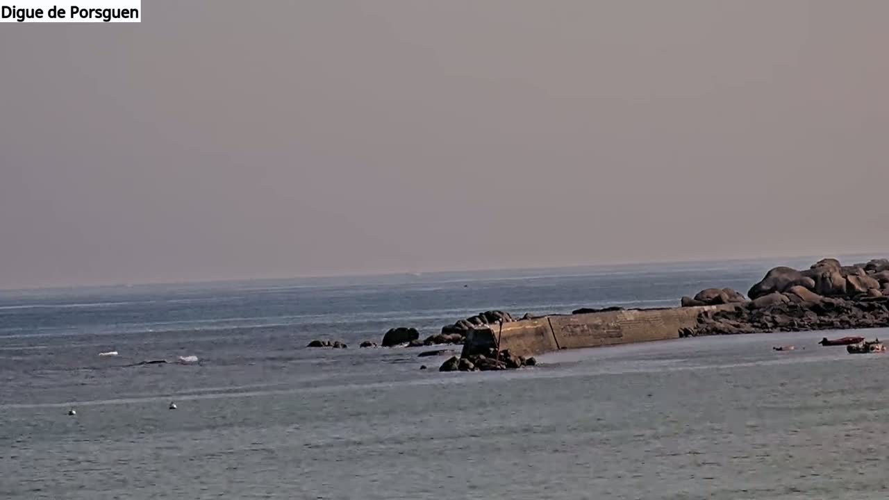 A hazy day reveals a rocky coastline with a low concrete wall extending into a calm sea, where a few small boats are visible.