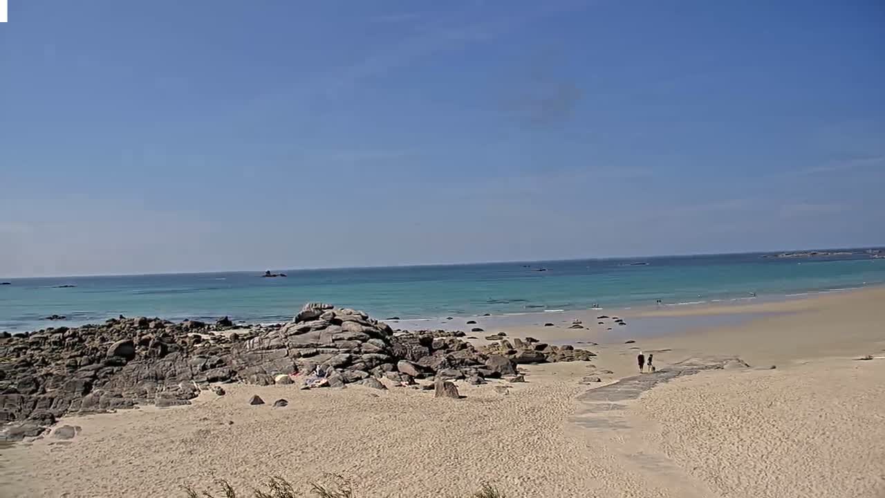 A sandy beach with rocky outcrops meets a calm, light teal ocean under a clear blue sky.