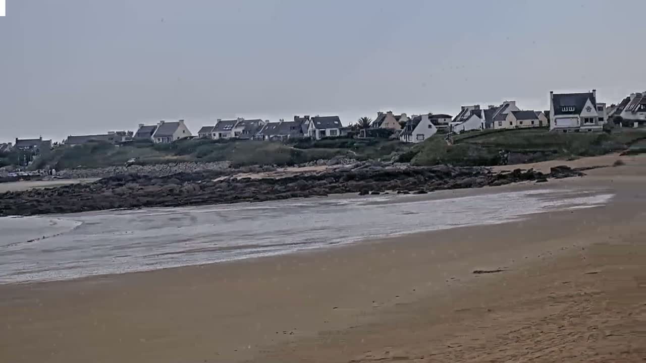 A sandy beach stretches towards a line of houses situated on a rocky outcrop overlooking the ocean under an overcast sky.