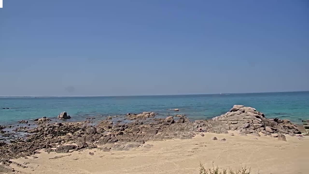 A sandy beach with dark rocks extends into a calm, turquoise ocean under a clear blue sky.