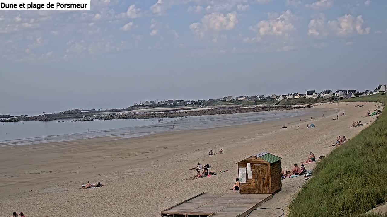 A sandy beach with scattered people sunbathing and swimming, under a partly cloudy sky, stretches out to a calm ocean and a distant shoreline with buildings.