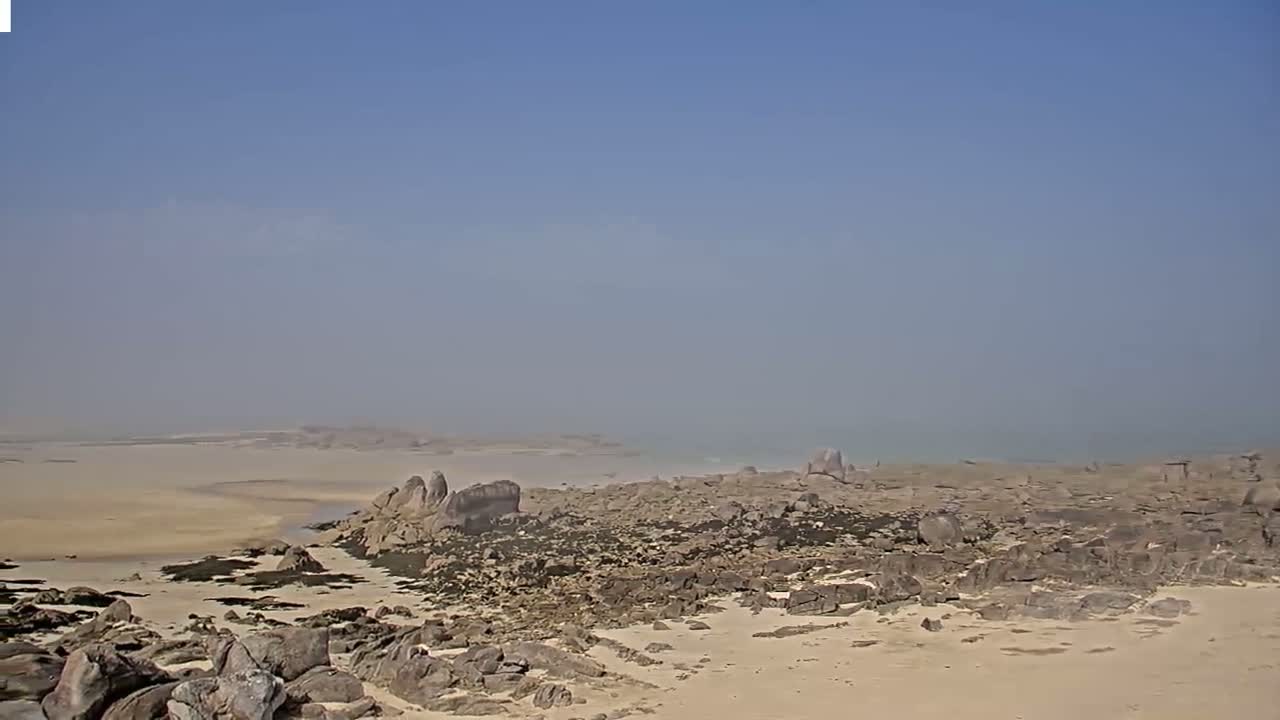 A hazy, sunny day reveals a rocky shoreline with a sandy beach, extending to a calm body of water in the distance.