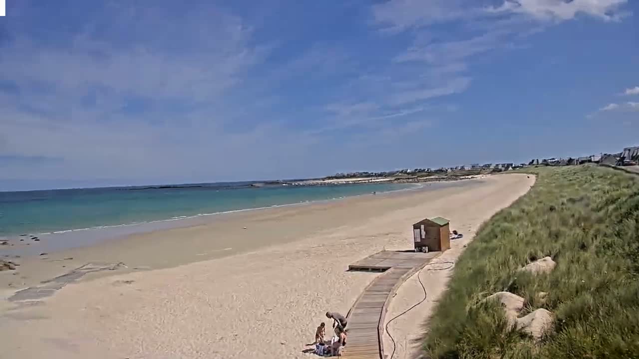 A sandy beach with calm, light-blue water stretches under a mostly sunny sky, with a few people near a wooden walkway and small structure on the shore.