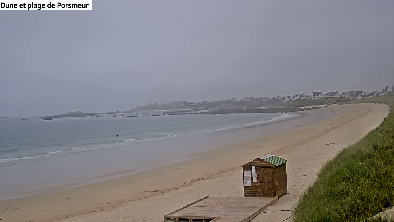 A sandy beach stretches out to a calm, hazy ocean under an overcast sky, with a small wooden structure near the shore and a distant village visible.