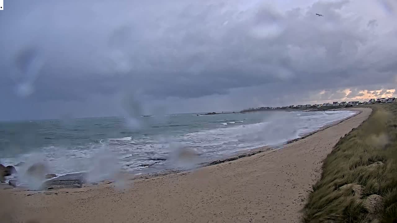 Large waves crash onto a sandy beach and rocky shore under a dark, heavily overcast sky with visible camera lens droplets, suggesting stormy and wet weather along a coastline featuring distant houses and a grassy dune.